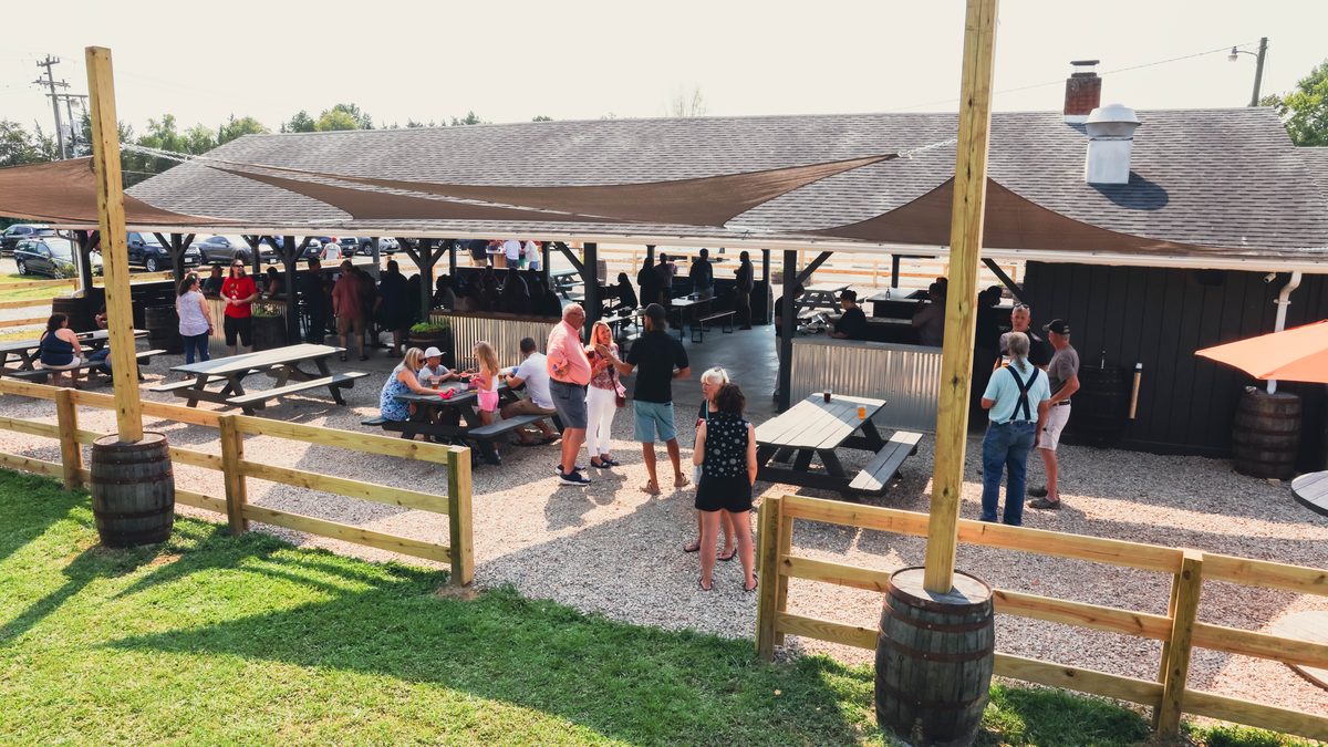 The gravel patio by the pavilion from above: groups standing and at picnic tables, a rail fence on barrels, shade sails, the lawn in front